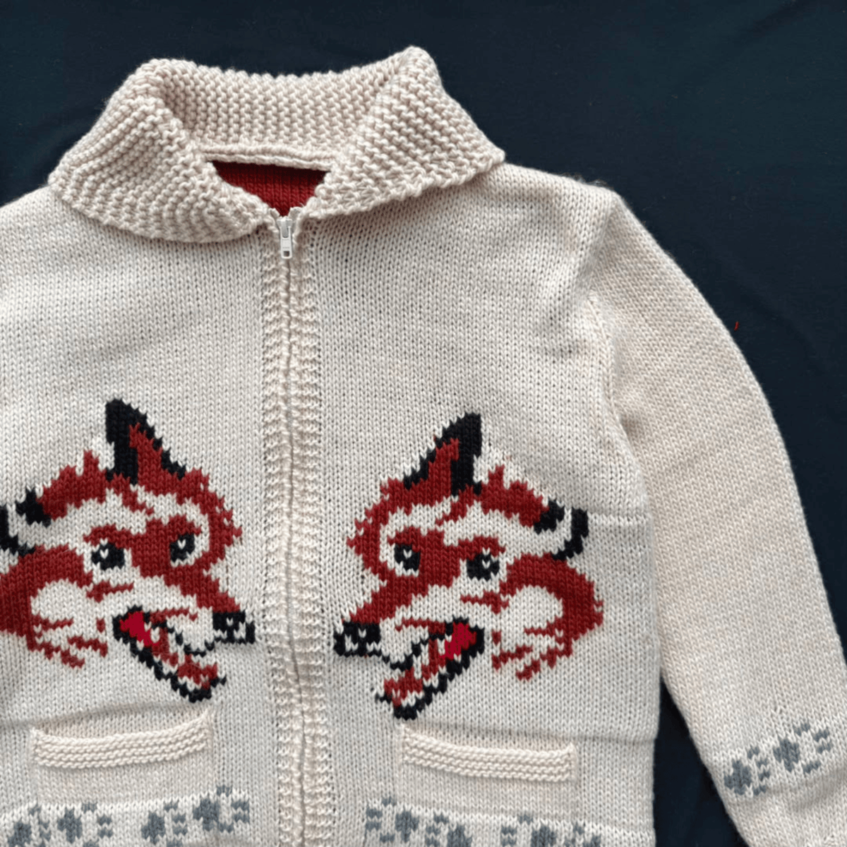 A cream-colored knit zip-up cardigan featuring two large red and black fox head motifs on the front, with a ribbed knit collar and front pockets, laid flat on a dark navy background.