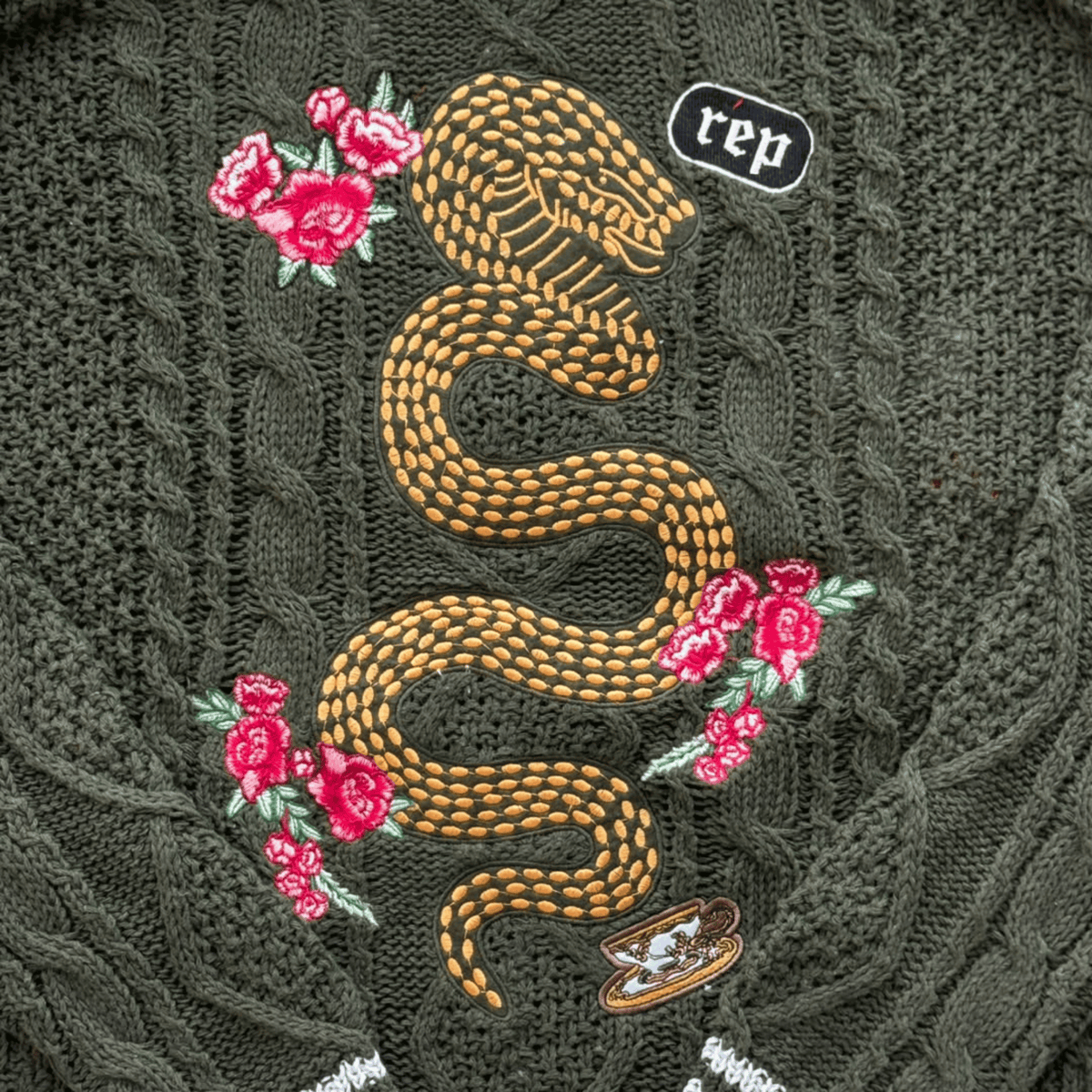Close-up of a dark olive green cable-knit sweater featuring a large embroidered gold snake, pink roses, and a small Rep logo patch.