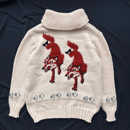 A cream-colored knit turtleneck sweater featuring two large red foxes knitted into the chest and small gray paw prints along the hem, laid flat on a dark navy background.