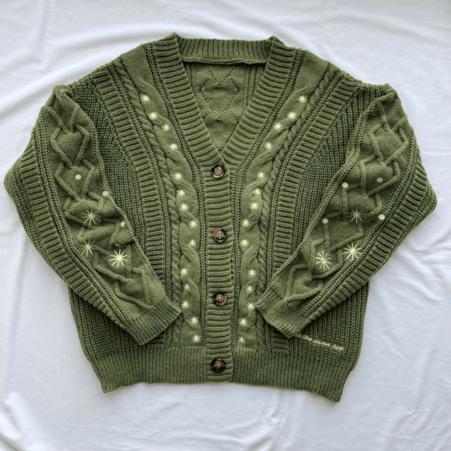 An olive green cable-knit cardigan sweater with cream-colored embroidered star and dot patterns, laid flat on a white fabric background.