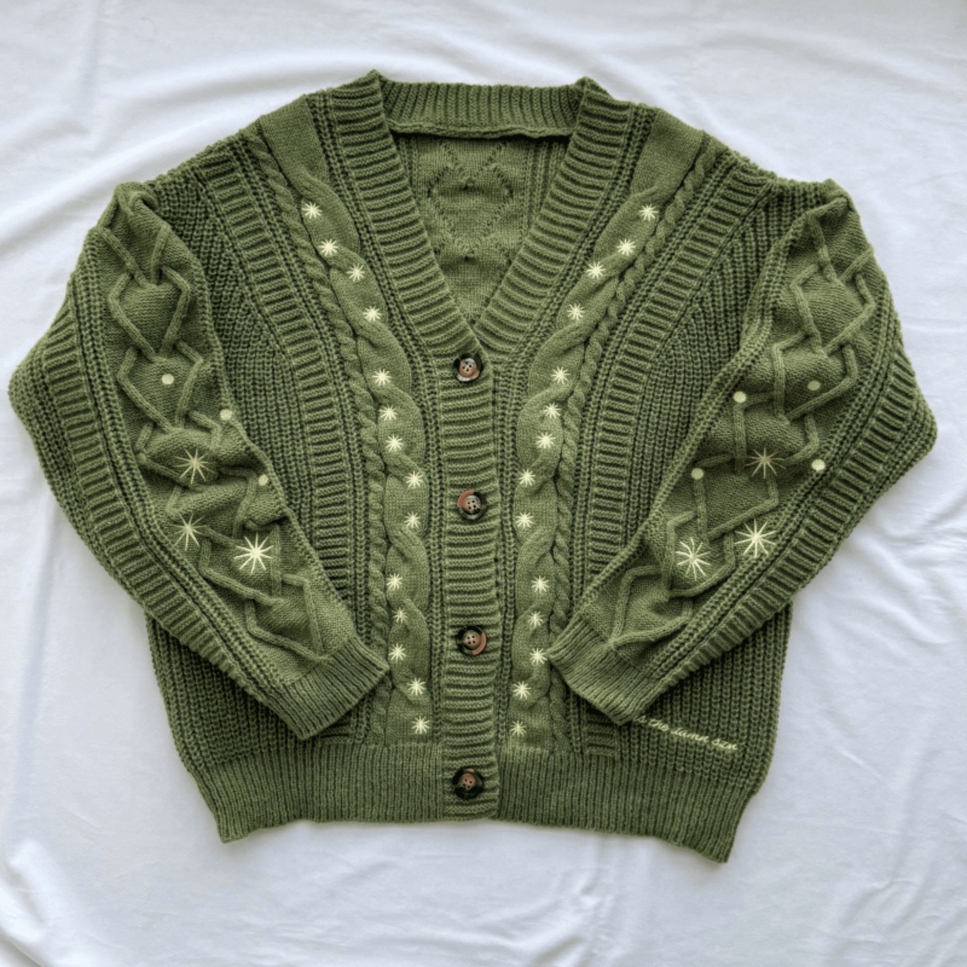 An olive green cable-knit cardigan sweater with cream-colored embroidered star and dot patterns, laid flat on a white fabric background.