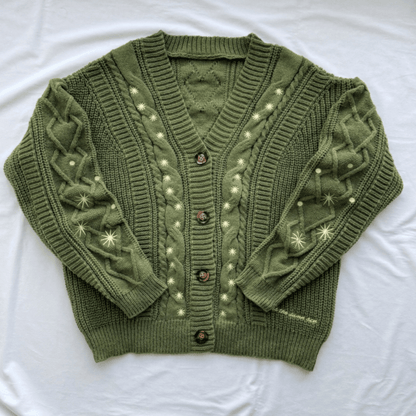 An olive green cable-knit cardigan sweater with cream-colored embroidered star and dot patterns, laid flat on a white fabric background.