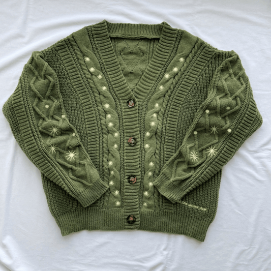 An olive green cable-knit cardigan sweater with cream-colored embroidered star and dot patterns, laid flat on a white fabric background.