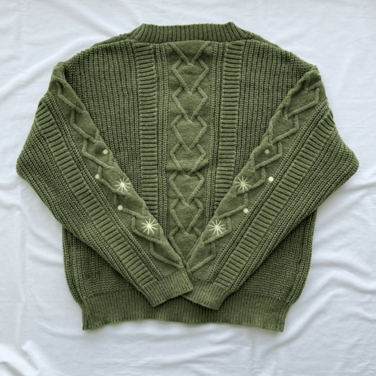 An olive green cable-knit sweater with decorative cream-colored embroidered star patterns on the sleeves, laid flat on a white fabric background.