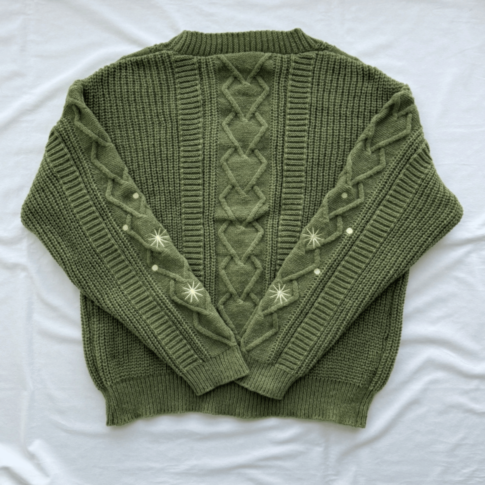 An olive green cable-knit sweater with decorative cream-colored embroidered star patterns on the sleeves, laid flat on a white fabric background.
