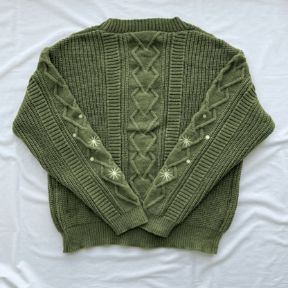 An olive green cable-knit sweater with decorative cream-colored embroidered star patterns on the sleeves, laid flat on a white fabric background.