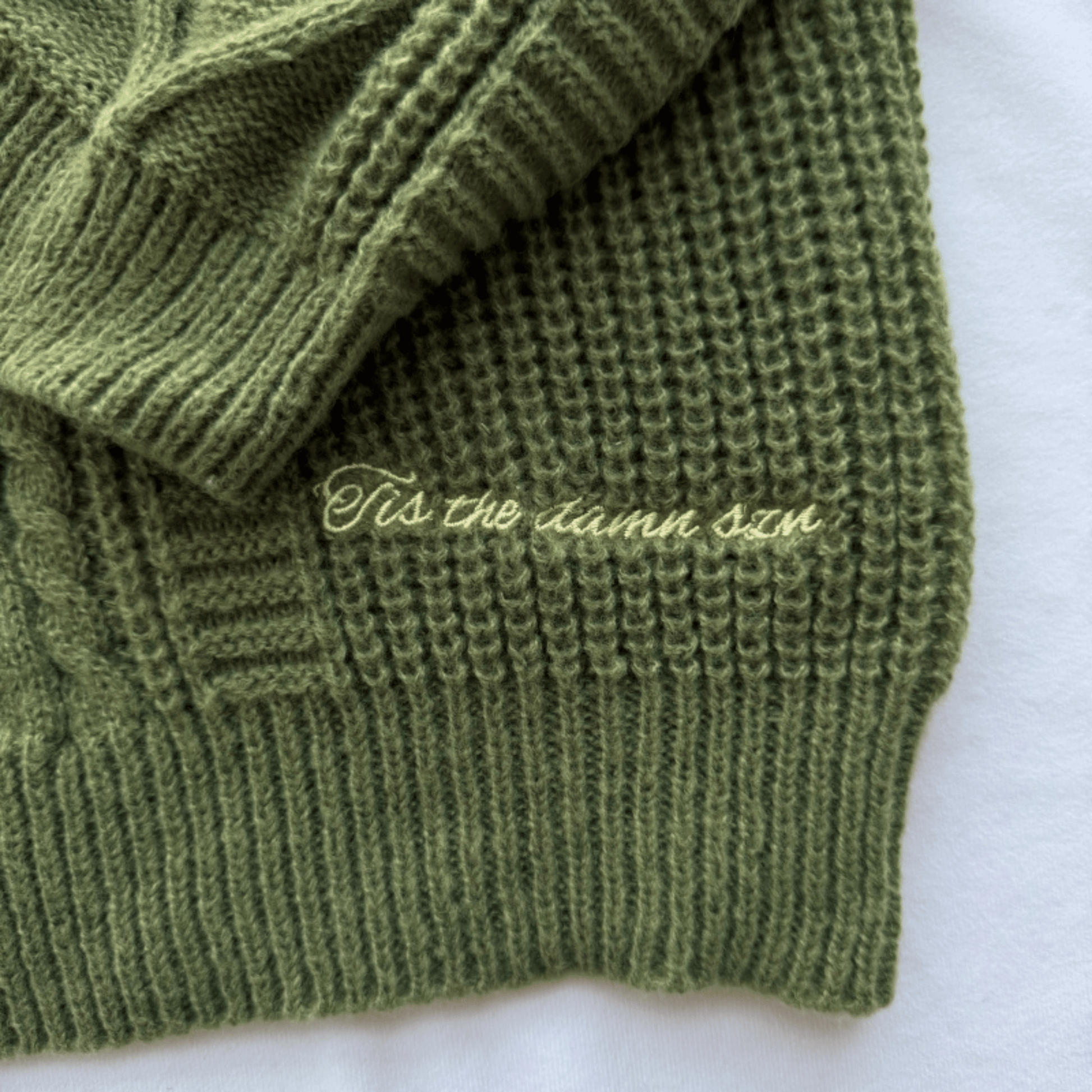 Close-up of an olive green ribbed knit sweater with cursive embroidery reading 'tis the damn szn' on a white background.