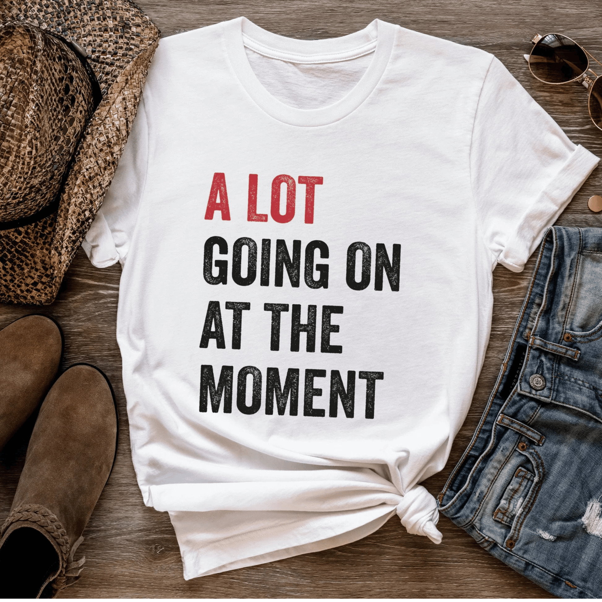 White t-shirt with text 'A LOT GOING ON AT THE MOMENT' on a wooden surface with shoes and denim.