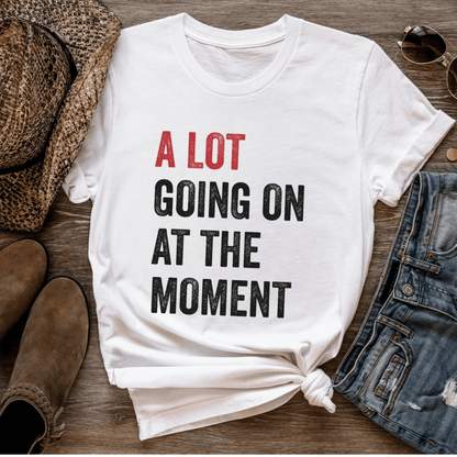 White t-shirt with text 'A LOT GOING ON AT THE MOMENT' on a wooden surface with shoes and denim.
