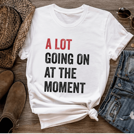 White t-shirt with text 'A LOT GOING ON AT THE MOMENT' on a wooden surface with shoes and denim.