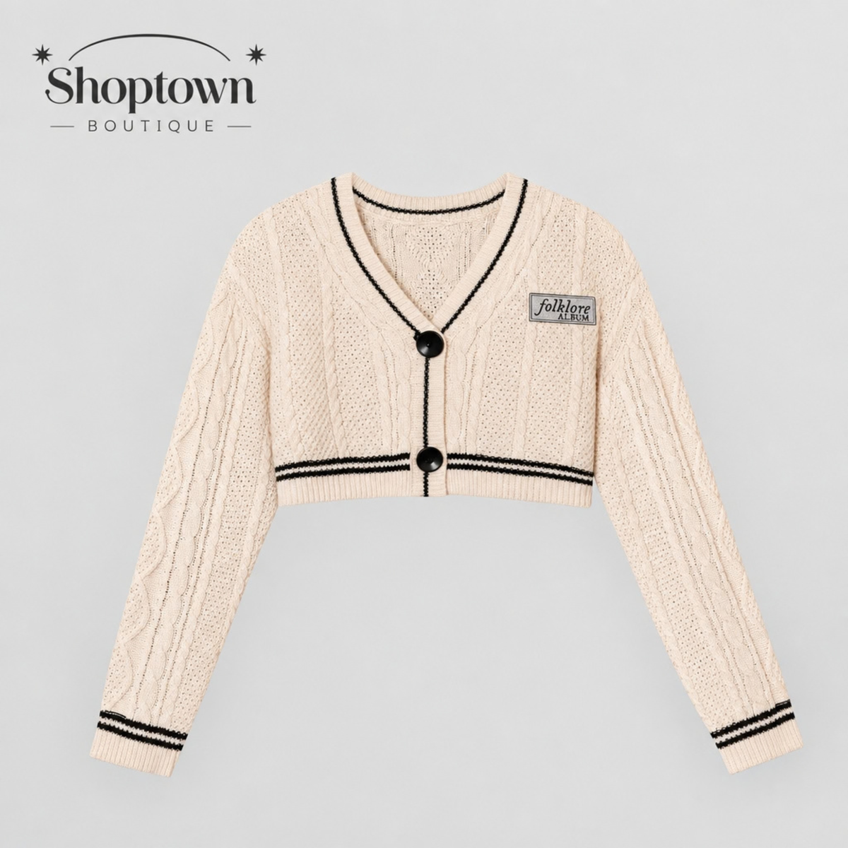 A cream-colored folklore knit cardigan sweater with black trim, black buttons, and a small 'fashionable' patch on the chest, displayed against a light gray background with the 'Shoptown Boutique' logo in the top left corner.