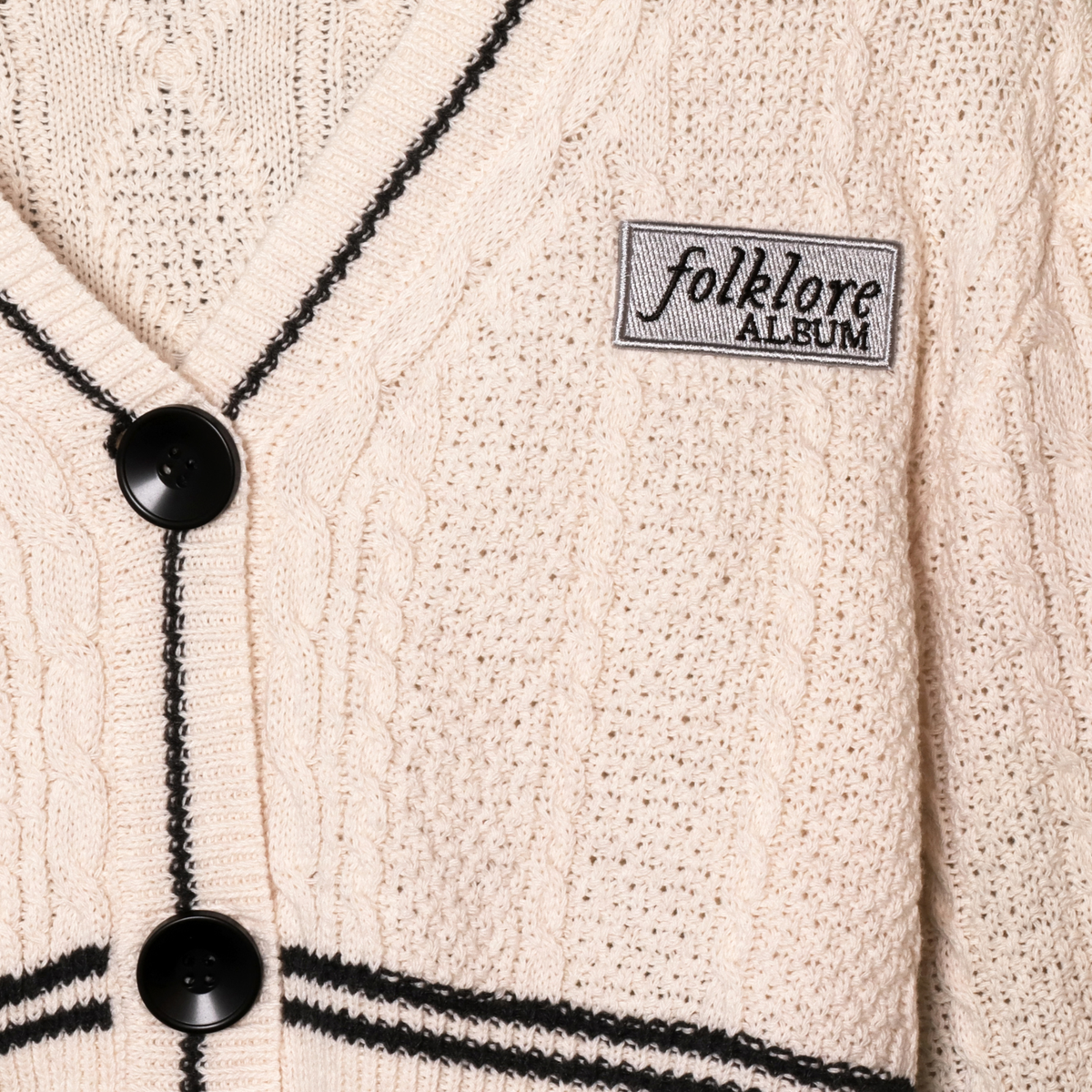 Close-up of a cream-colored folklore knit cardigan sweater with black trim and black buttons, featuring a woven label that reads 'folklore ALBUM'.