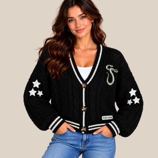 A woman wearing a black cable-knit cardigan with white trim, white embroidered stars on the sleeves, a green snake brooch on the chest, and gold-tone buttons, paired with blue jeans against a beige studio background.