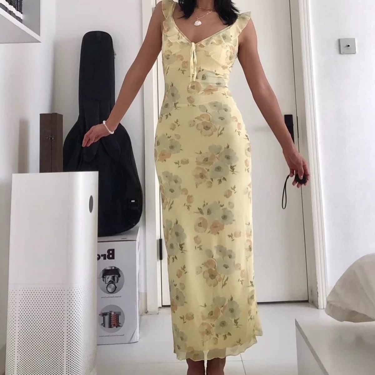 Woman in a yellow floral dress standing in a room with white walls and minimal furniture.