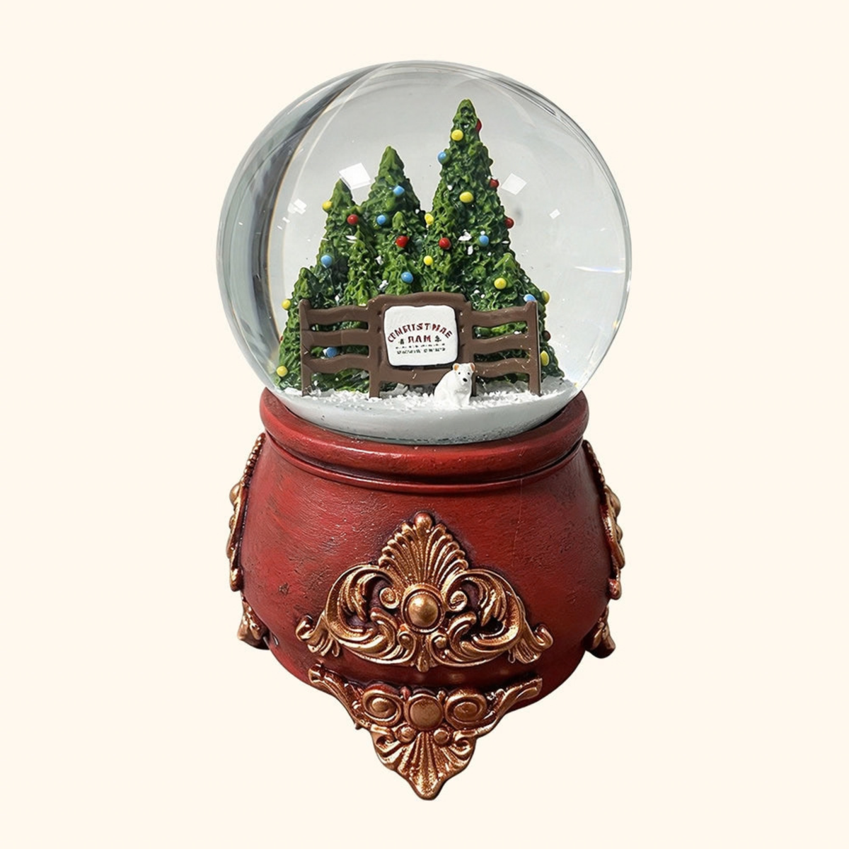 A decorative snow globe figurine with a red base and ornate gold detailing, featuring miniature Christmas trees, a fence, a small sign reading 'Christmas Tree Farm', and white faux snow inside a clear globe, shown on a plain light beige background.