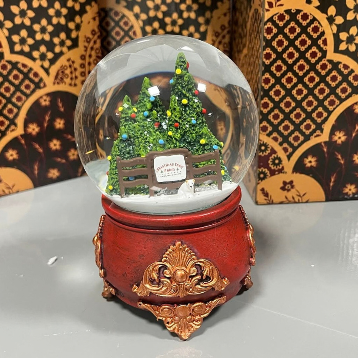 A decorative red and gold snow globe featuring miniature Christmas trees, a fence, and a small sign that reads'Christmas Tree Farm' displayed on a light gray surface with patterned boxes in the background.