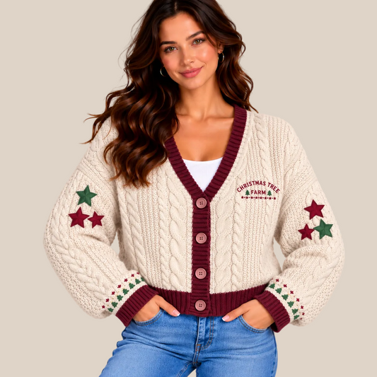 A woman wearing a cream cable-knit cardigan with burgundy trim, green and burgundy star details, and 'Christmas Tree Farm' embroidered on the chest, paired with blue jeans against a warm beige background.