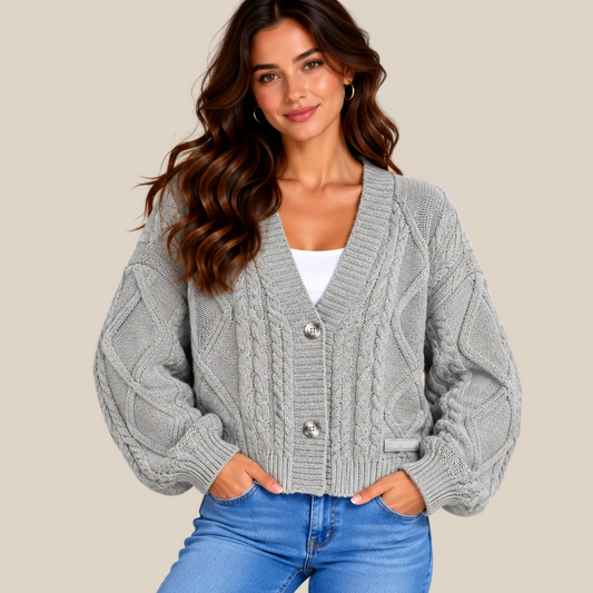 A woman wearing a light gray cable-knit cardigan with silver buttons over a white top and blue jeans, standing against a solid beige background.