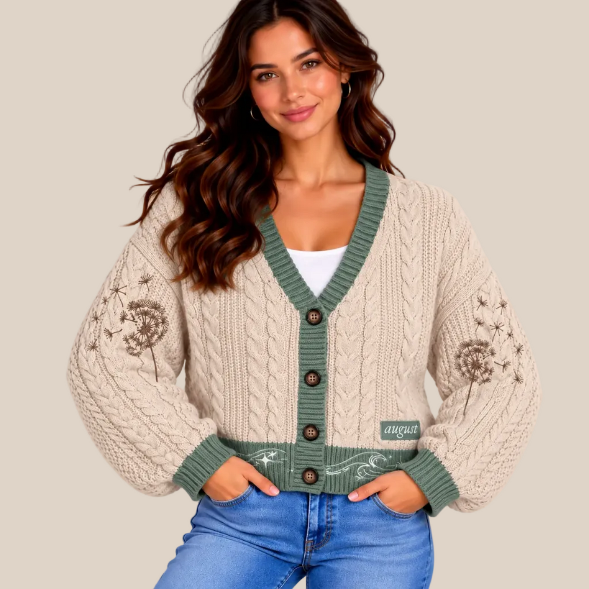 A woman wearing a cream cable-knit cardigan with sage green trim and button details, styled with blue jeans against a warm beige studio background.