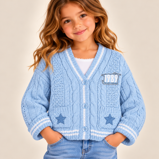 A young girl with wavy light brown hair smiling, wearing a light blue cable-knit cardigan with white stripes, star details, and a '1989' patch, paired with blue jeans against a solid beige background.
