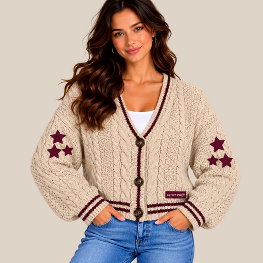 A woman wearing a cream cable-knit cardigan with burgundy trim and star details on the sleeves, paired with blue jeans, against a solid beige background.