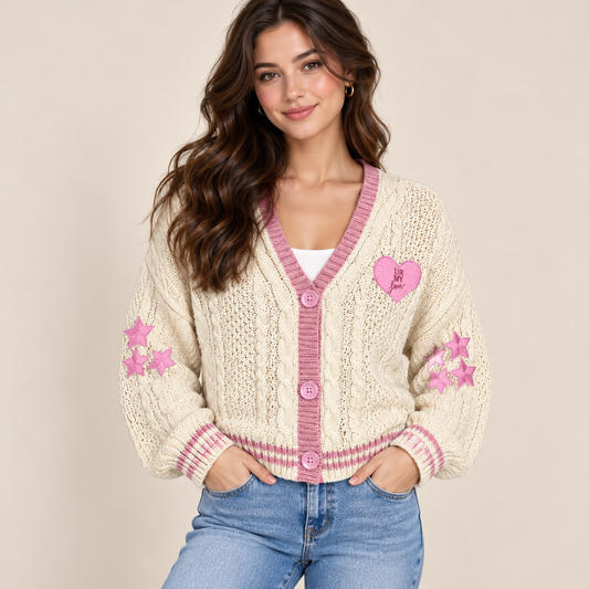 A woman wearing a cream cable-knit cardigan with pink trim, pink buttons, and pink embroidered stars and a heart on the sleeves, paired with blue jeans against a solid beige background.