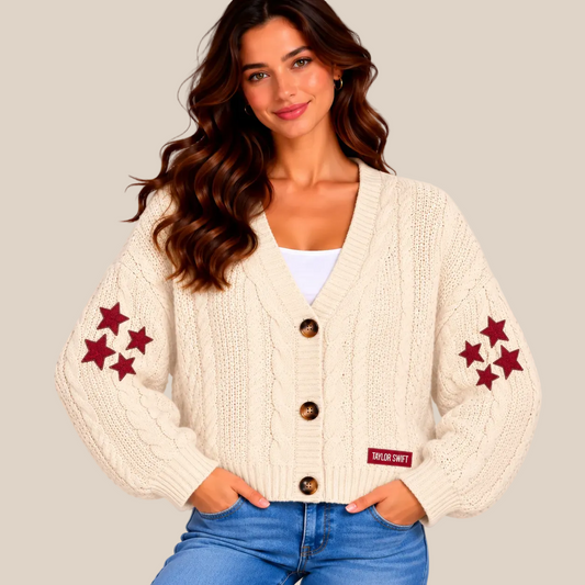 A woman wearing a cream cable-knit cardigan with red embroidered star clusters on the sleeves and a small red brand patch near the hem, paired with blue jeans against a soft beige studio background.