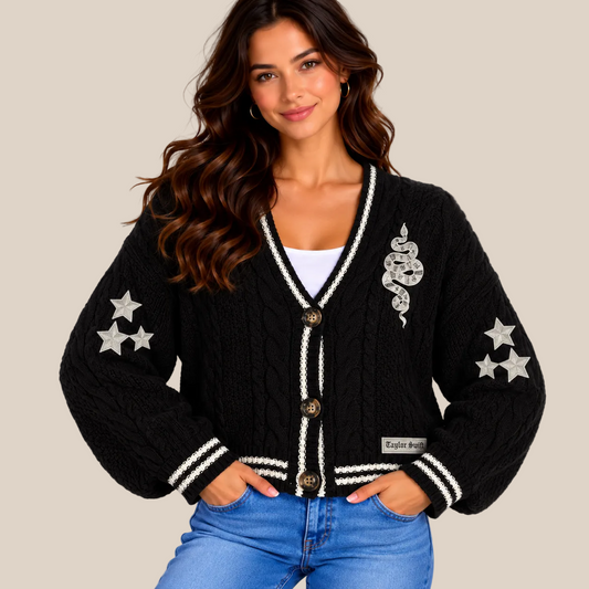 A woman wearing a black cable-knit cardigan with white striped trim, silver star patches on the sleeves, and a silver snake patch on the chest, paired with blue jeans against a beige studio background.