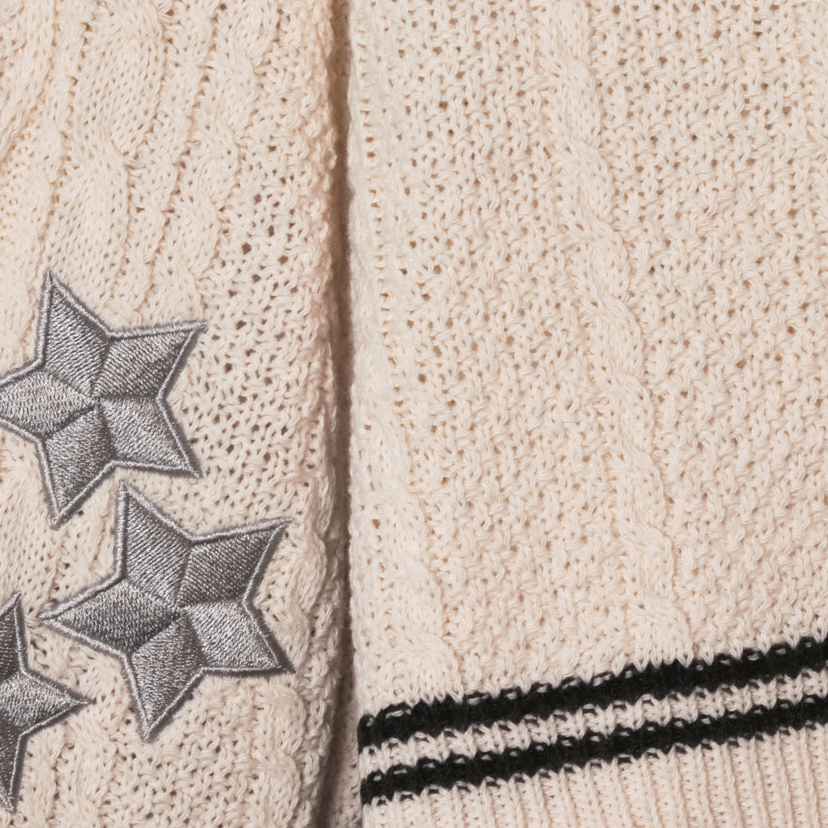 Close-up of a cream-colored folklore knit sweater showing the texture, a black double stripe at the hem, and gray embroidered stars on the sleeve.