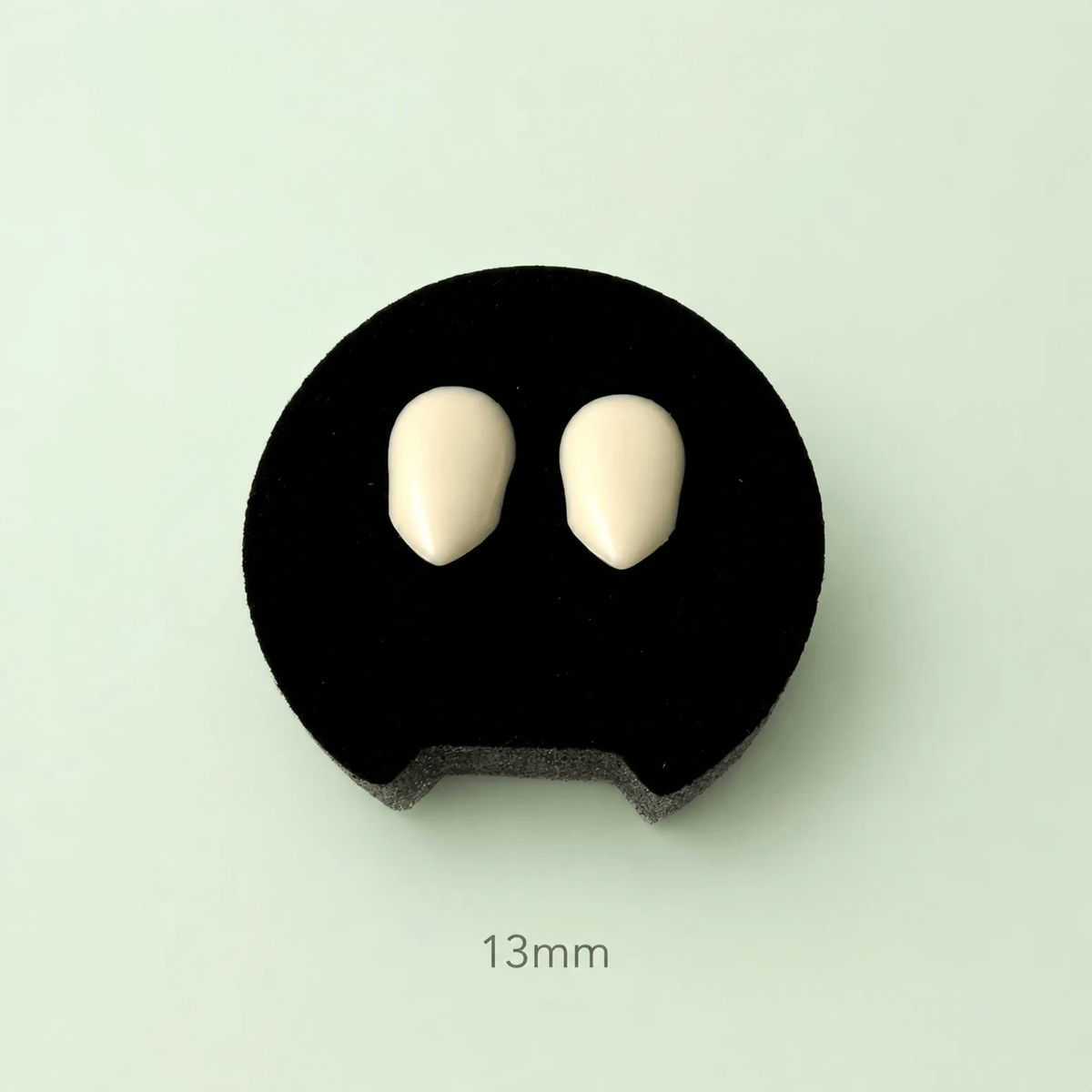 Two small off-white oval tooth-shaped pieces displayed on a black circular foam stand against a pale mint background, with the text '13mm' below.