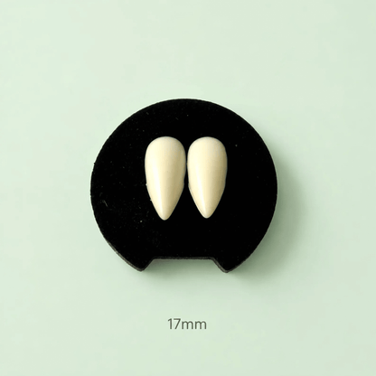 A pair of off-white pointed vampire fang nail art tips displayed on a black circular stand against a pale mint green background, with '17mm' text printed below.