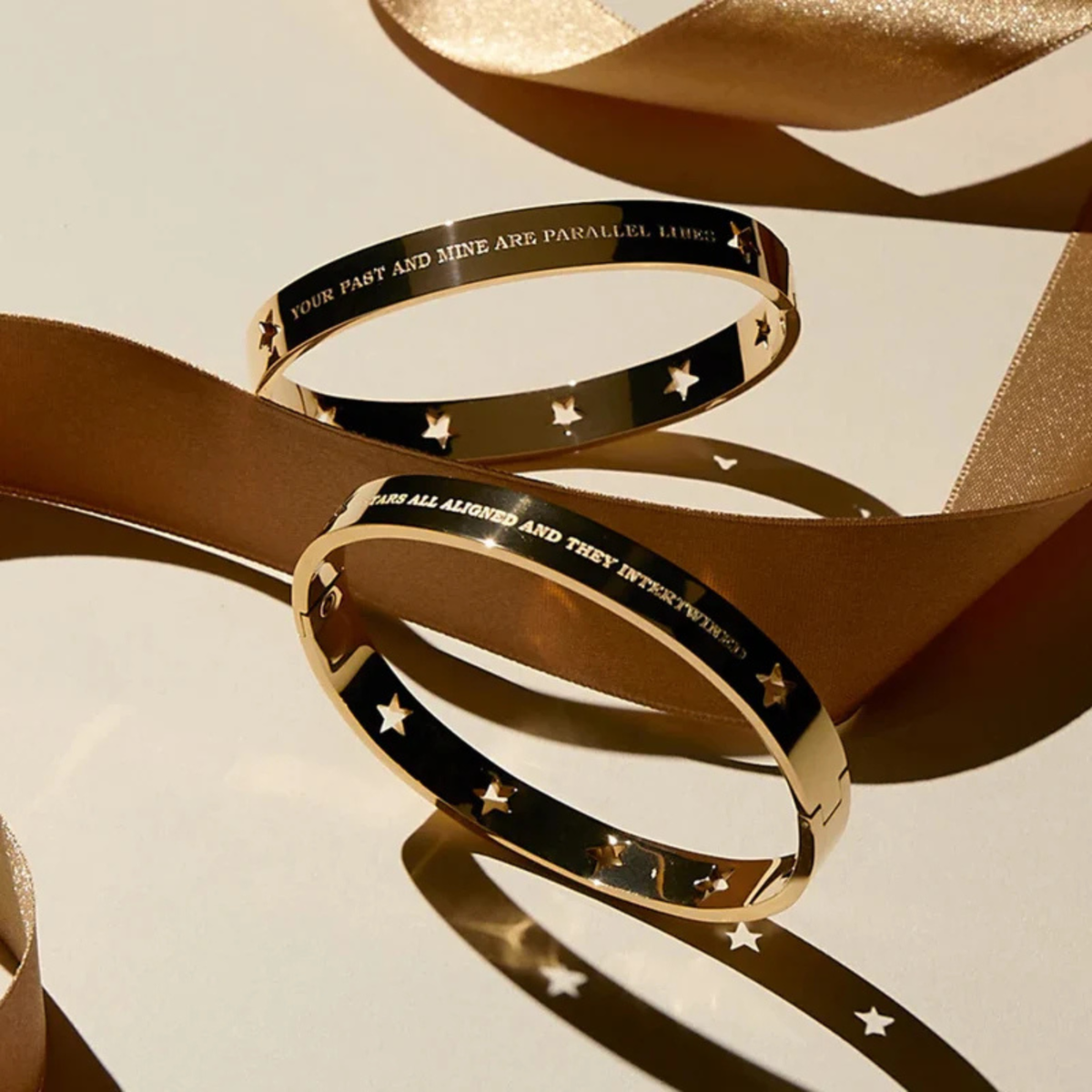 Two gold bangle bracelets with black inlay and engraved text, styled with a flowing champagne-colored ribbon on a light beige background.