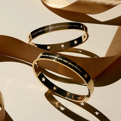 Two gold bangle bracelets with black inlay and engraved text, styled with a flowing champagne-colored ribbon on a light beige background.