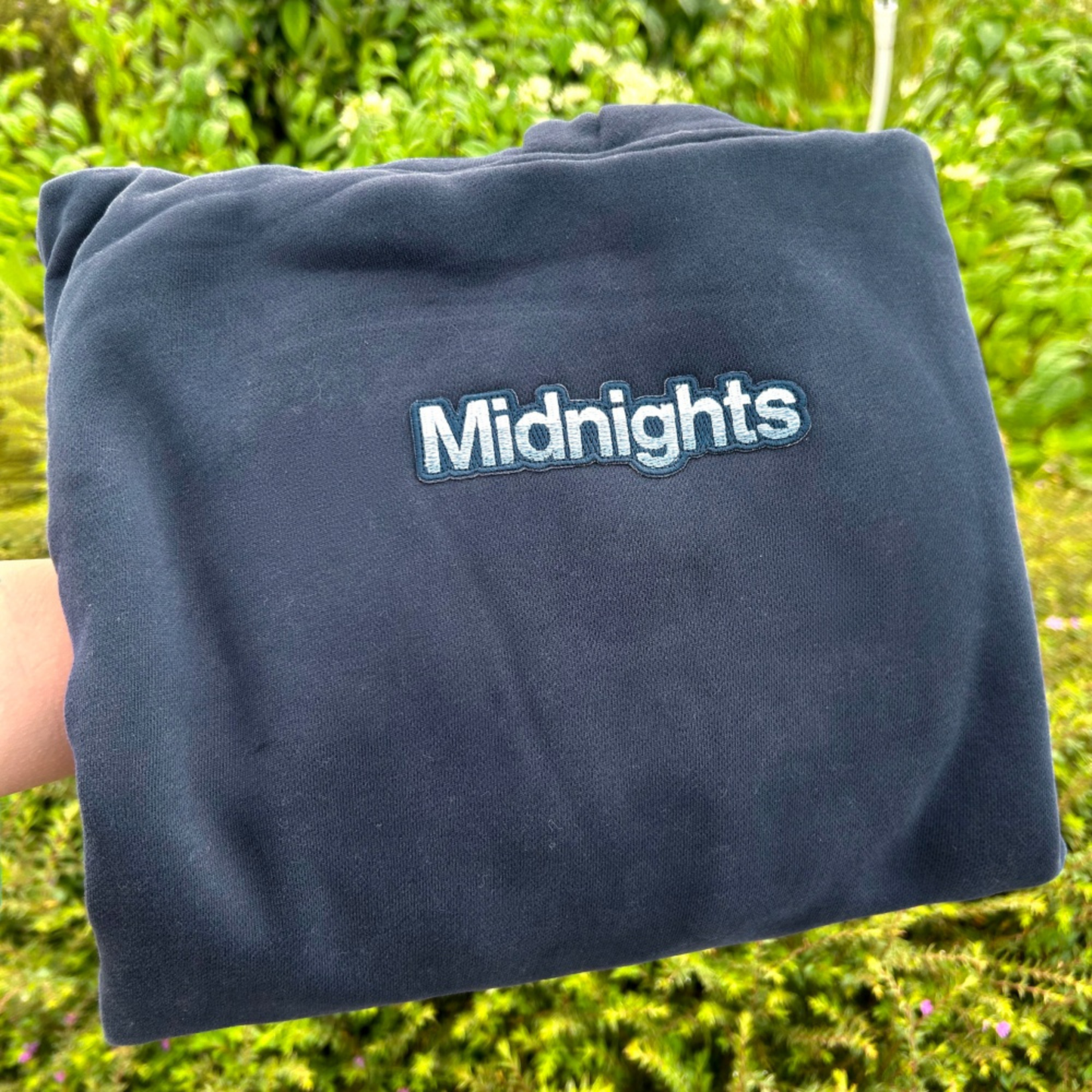 A folded navy blue sweatshirt or hoodie with the word 'Midnights' embroidered in white with a blue outline, held up by a hand against a blurred green outdoor background.