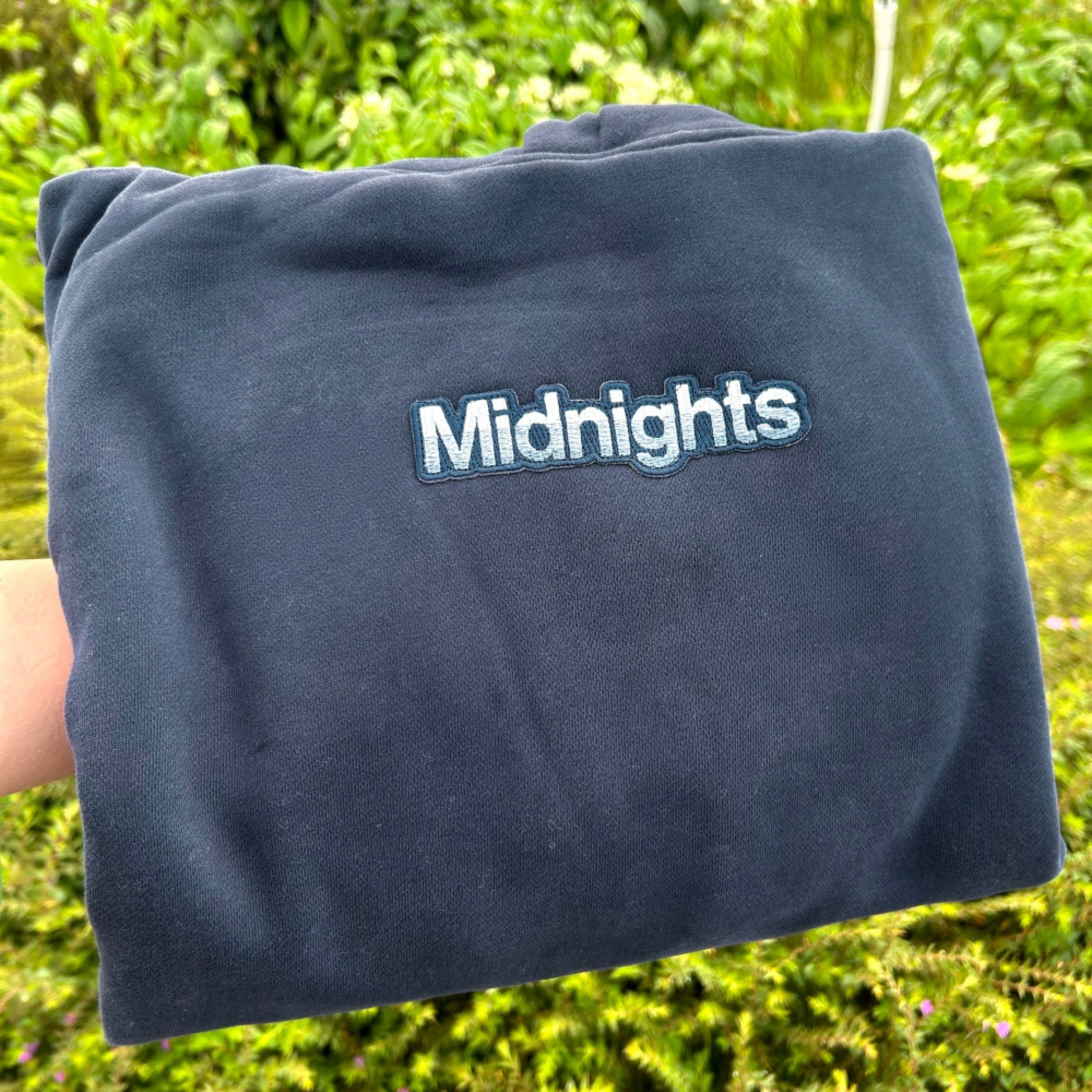 A folded navy blue sweatshirt or hoodie with the word 'Midnights' embroidered in white with a blue outline, held up by a hand against a blurred green outdoor background.