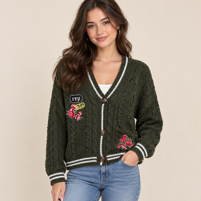 A woman wearing an olive green cable-knit cardigan with white striped trim, brown buttons, and colorful floral patches, paired with blue jeans against a beige background.