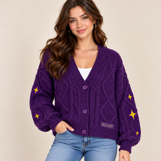 A woman wearing a purple cable-knit cardigan with yellow star details on the sleeves, paired with blue jeans and a white top, against a solid beige background.