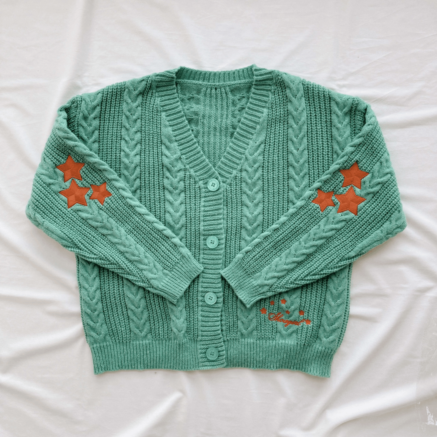 A mint green cable-knit cardigan sweater with orange embroidered star accents on the sleeves and lower front, laid flat on a white fabric background.
