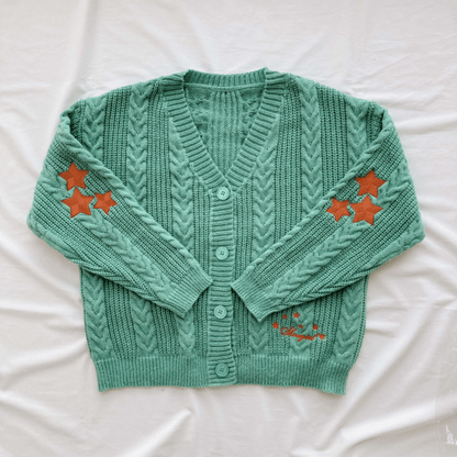 A mint green cable-knit cardigan sweater with orange embroidered star accents on the sleeves and lower front, laid flat on a white fabric background.