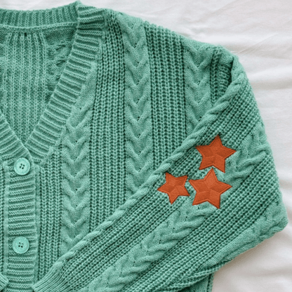 A mint green cable-knit cardigan sweater laid flat on a white fabric background, featuring three orange embroidered stars on the sleeve and matching mint buttons on the front.