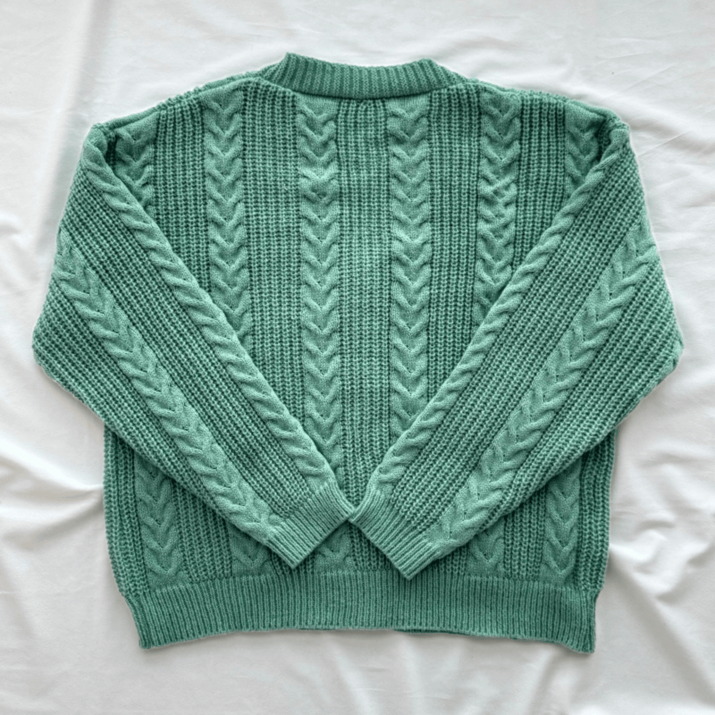 A sage green cable-knit sweater with a crew neck and long sleeves, laid flat on a white fabric background.