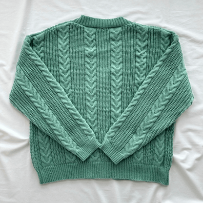 A sage green cable-knit sweater with a crew neck and long sleeves, laid flat on a white fabric background.