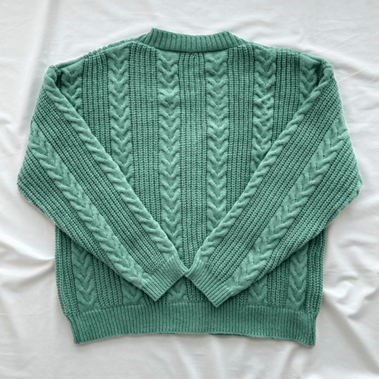 A sage green cable-knit sweater with a crew neck and long sleeves, laid flat on a white fabric background.