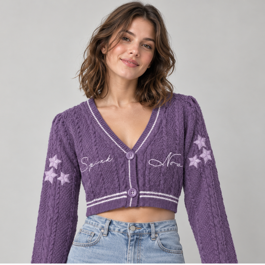 A woman wearing a purple cable-knit cropped cardigan with white embroidered script and star details, paired with light blue jeans, against a plain light gray background.