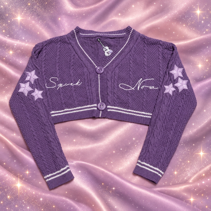 A purple cable-knit cropped cardigan sweater with white striped trim, featuring 'Spark' and 'Nora' embroidered in white script on the chest, and three lavender stars on each sleeve. The cardigan is laid flat on a light gray tiled floor.