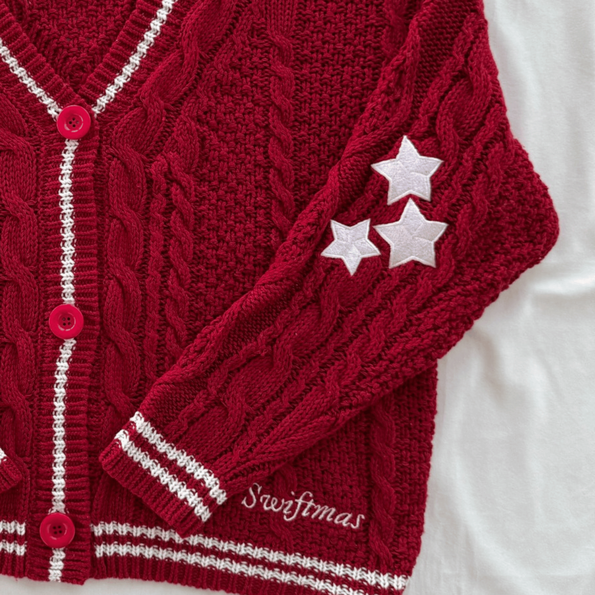 A close-up of a deep red cable-knit cardigan with white striped trim, pink buttons, and three white embroidered stars on the sleeve. The word 'Swiftmas' is embroidered near the cuff, and the cardigan is laid flat on a white fabric background.