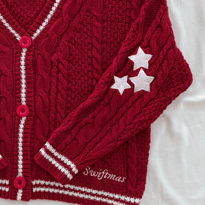 A close-up of a deep red cable-knit cardigan with white striped trim, pink buttons, and three white embroidered stars on the sleeve. The word 'Swiftmas' is embroidered near the cuff, and the cardigan is laid flat on a white fabric background.