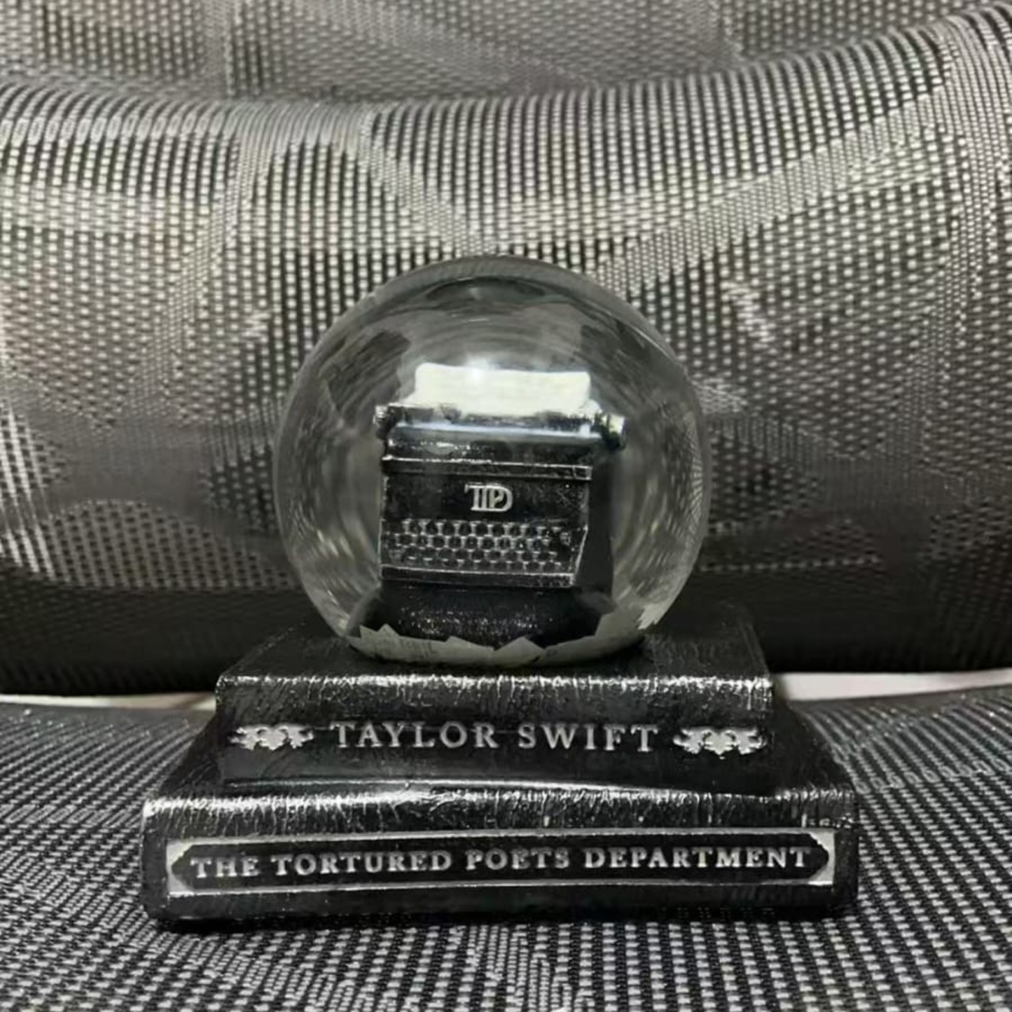 Crystal ball with a small keyboard inside on a stand labeled 'Taylor Swift The Tortured Poets Department' against a textured black background.