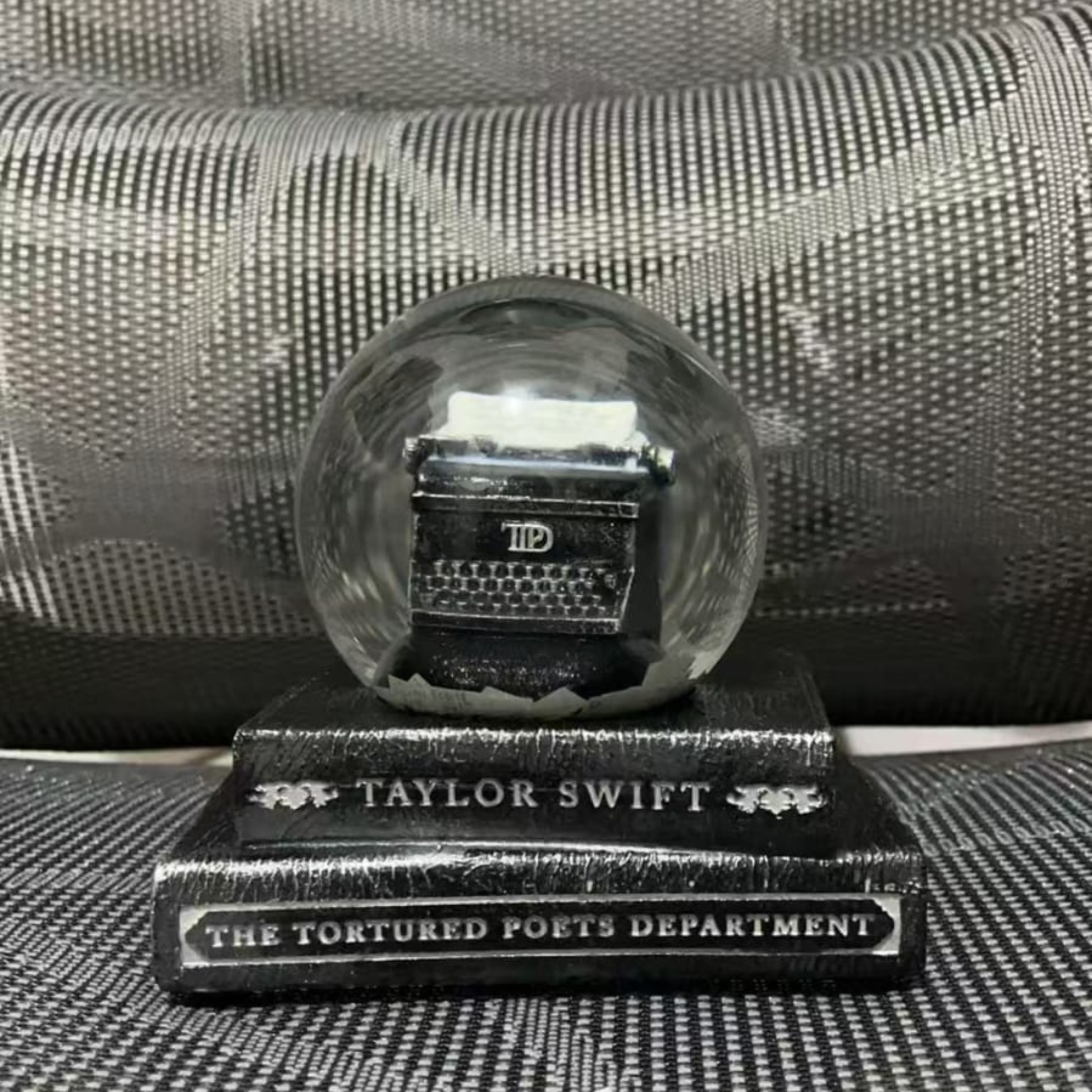 Crystal ball with a small keyboard inside on a stand labeled 'Taylor Swift The Tortured Poets Department' against a textured black background.