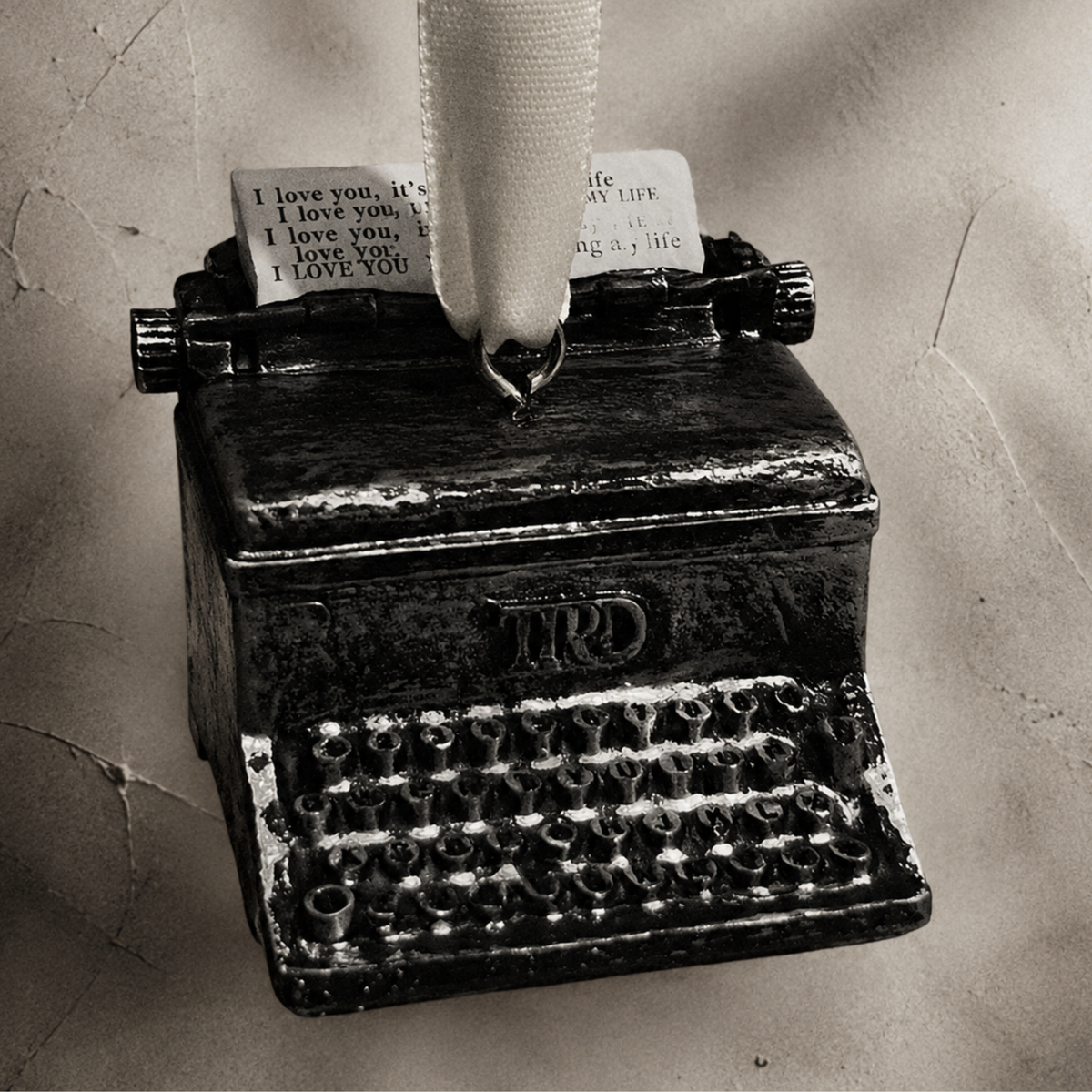 A vintage-style TTPD typewriter ornament or decorative object with a ribbon loop at the top and a small printed note, shown against a textured beige background.